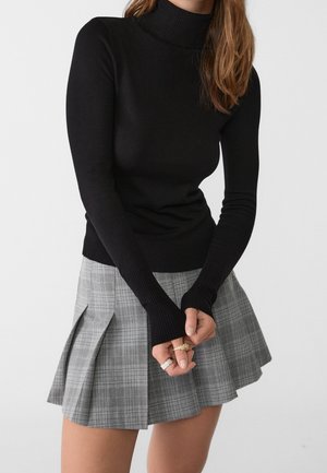 Femme portant un pull col roulé noir à manches longues et une mini-jupe plissée à carreaux grise, tenant les mains devant elle.
