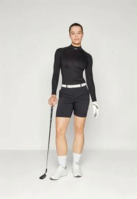 Melnas garās piedurknes golfa krekls, kombinēts ar melniem šortiem, baltu jostu un baltiem apaviem, tur rokās golfa nūju. Plūdena auduma un sportisks piegriezums.