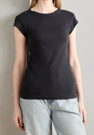 Kobieta ubrana w dopasowany czarny krótki rękaw i jasnoniebieskie dżinsy na jednolitym tle.