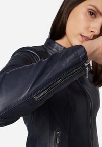 Marinblå läderjacka med dragkedja, dragkedjeslutningar på ärmarna och texturerade axeldetaljer. Slät textur med detaljerad sömnadsaccent.
