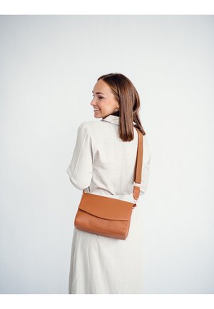 Kladěná kůže hnědá crossbody taška s klopou, nastavitelným popruhem a embosovanými detaily, nošená modelkou v svetle barevném šatu.