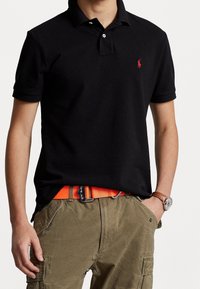 Svart polo skjorta med en liten röd broderad logotyp, parat med khakifärgade cargobyxor och ett orange bälte. Har korta ärmar och krage.