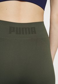 Zbliżenie na talię osoby, ubraną w ciemnozielone legginsy bezszwowe z wysokim stanem, na którym znajduje się logo Puma w ciemnym druku.