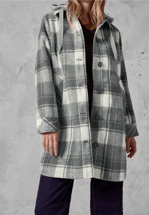 Kvinde iført en grå og hvid ternet knap-op frakke med hætte og forlommer, kombineret med mørke bukser mod en grå tekstureret baggrund.