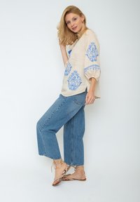 Beige linnen blouse met blauwe bloemenpatronen, gecombineerd met lichtblauwe, hoge taille wijde jeans. Draagt bij beige sandalen met bandjes.