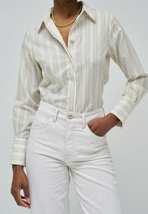 Personne portant une chemise à rayures beige boutonnée, rentrée dans un pantalon blanc taille haute, main droite dans la poche et un collier chaîne en or.