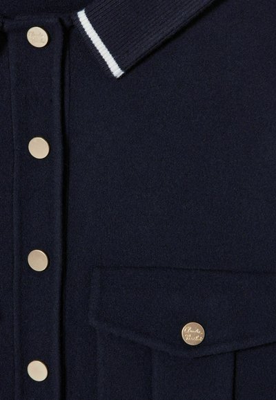 Polo de lana navy con cuello a rayas blancas, botones dorados y un bolsillo en el pecho con un acento dorado de la marca.