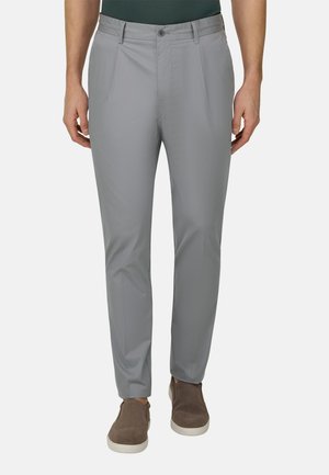 Mand iført lysegrå slim-fit bukser og brune slip-on sko, stående med armene afslappet langs siden mod en ensfarvet baggrund.