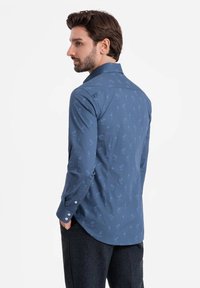 Camisa de manga larga para hombre en algodón de color azul oscuro con un sutil estampado de palmeras, cuello tradicional y puños abotonados. Textura suave, diseño ajustado.