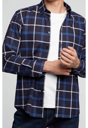 Mann trägt ein dunkelblau kariertes Hemd mit blauen und weißen Linien über einem weißen T-Shirt und richtet seinen Ärmel.