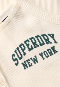 Crèmekleurige ribstof met groene geborduurde tekst "SUPERDRY NEW YORK." Kenmerkt zich door een ronde halslijn en een knoopdetail aan de zijkant.