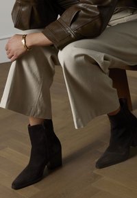 Schwarze Wildleder-Stiefeletten mit spitzer Zehenpartie und grobem Absatz, kombiniert mit hellbeigen, weit geschnittenen Hosen und einer braunen Lederjacke.