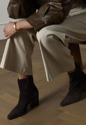 Schwarze Wildleder-Stiefeletten mit spitzer Zehenpartie und grobem Absatz, kombiniert mit hellbeigen, weit geschnittenen Hosen und einer braunen Lederjacke.