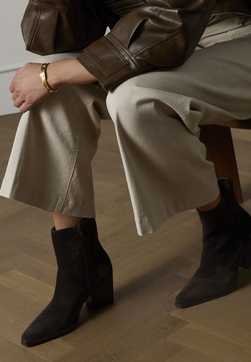 Schwarze Wildleder-Stiefeletten mit spitzer Zehenpartie und grobem Absatz, kombiniert mit hellbeigen, weit geschnittenen Hosen und einer braunen Lederjacke.