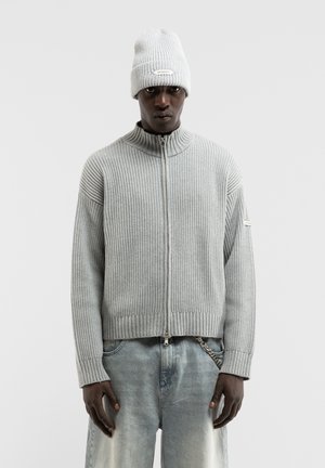Mann trägt hellgrauen, gerippten Zip-up-Pullover, passende gestrickte Mütze und ausgewaschene blaue Jeans mit Kettendetail, steht vor schlichten Hintergrund.