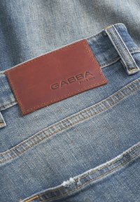 Kék színű denim farmernadrág, barom bőr címkével, amelyen a "GABBA" felirat és az "1983" évszám látható. Figyelemre méltó varrási részletek is láthatók.