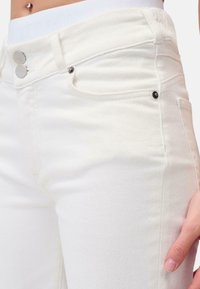 Jeans in denim bianco con vestibilità slim, caratterizzati da bottoni in metallo argentato, design a cinque tasche e dettagli di cucitura minimi. Texture liscia.