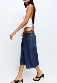 Culottes en jean bleu foncé, présentant une coupe jambes larges, complétées par une ceinture marron à clous argentés. Portées avec un débardeur blanc et des talons.