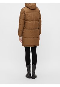 Bruine pufferjas met capuchon, met horizontale gestikte secties, lange mouwen en een knielengte ontwerp. Gdragen over zwarte leggings en laarzen.