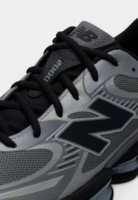 Chaussure de sport grise avec des accents noirs, dotée d'une tige en mesh texturé, d'une languette rembourrée et d'un logo contrastant, maintenue par des lacets noirs.