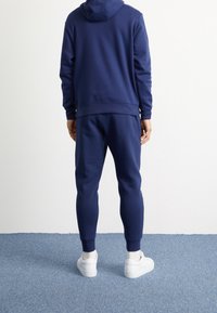 Sudadera azul marino y joggers a juego, hechos de tela suave. Detalle de capucha, puños ajustados y cinturilla elástica. Zapatillas blancas.