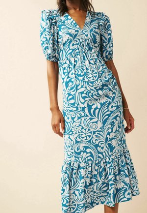 Femme portant une robe midi à motifs floraux bleus et blancs avec des manches bouffantes et un ourlet à volants sur un fond uni.