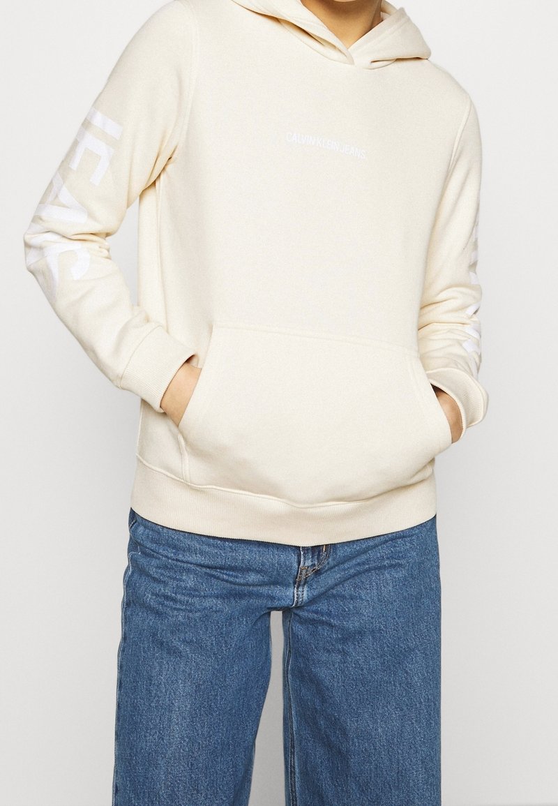 Persona che indossa un cappuccio crema di Calvin Klein con tasca a marsupio e jeans blu, con le mani infilate nelle tasche del cappuccio, torso visibile.