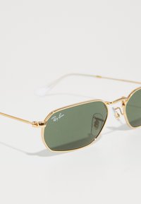 Gafas de sol de metal dorado con lentes verdes, con forma octagonal y un marco de alambre delgado. Logo de Ray-Ban visible en una lente.