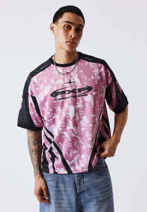 Joven con una camiseta rosa y negra PXG "Change the Game" y vaqueros azules, de pie frente a un fondo claro y liso.
