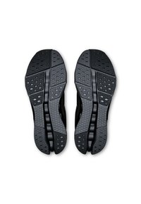 Semelles de chaussures de sport noires avec des motifs texturés, des grips circulaires et en forme de diamant, et des rainures centrales pour la flexibilité.