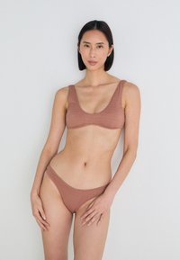 Ruda įripiama bikini komplektas, kuriame yra liemenėlė su apvalia iškirpte ir žemai sėdinčios thong tipo kelnaitės, abi pagamintos iš tekstūruoto audinio. Paprastas dizainas, be puošmenų.