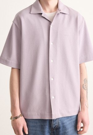 Mann trägt ein hellviolettes kurzärmeliges Hemd mit Knöpfen, weißes Unterhemd, blaue Jeans, Armband am linken Handgelenk und Tätowierung am rechten Arm.