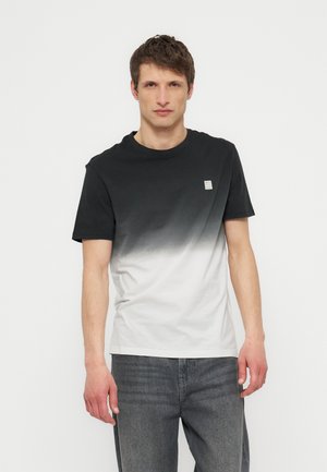 Junger Mann trägt ein schwarz-weiß verlaufendes T-Shirt und graue Jeans und steht vor einem schlichten weißen Hintergrund.