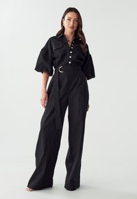 Chemise noire à manches bouffantes, avec une ceinture à boucle circulaire et un pantalon à jambes larges. Tissu lisse, design minimaliste.