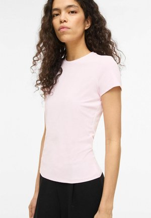 Femeie cu păr lung și creț, purtând un tricou roz deschis, cu mânecă scurtă, mulat, și pantaloni negri, stând pe un fundal alb simplu.