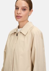 Giacca beige con un ampio colletto, chiusura con bottoni e una texture liscia. Il design presenta una vestibilità rilassata e dettagli di cucitura discreti.