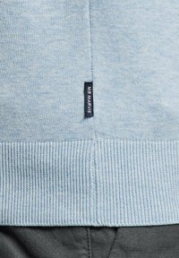 Ourlet de pull tricoté bleu clair avec une petite étiquette noire portant l'inscription "MR MARVIS" au-dessus d'un pantalon gris foncé.
