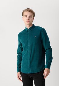 Camisa de pana en verde azulado con cuello abotonado, mangas largas y un pequeño parche de logo en el pecho. Presenta un corte recto y una tela texturizada.