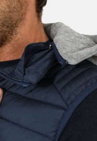 Gilet imbottito blu navy con cappuccio in tessuto grigio, caratterizzato da una texture liscia e chiusura zip visibile vicino al colletto.