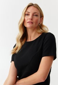 Robe noire à manches courtes avec un col rond et des coutures structurées. Tissu lisse et silhouette cintrée ; le modèle porte des boucles d'oreilles en or.