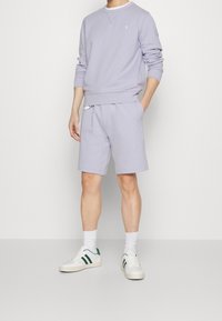 Ljuslila sweatshirt och matchande shorts i mjukt tyg, med en liten logotyp, vita strumpor och vita sneakers med gröna detaljer.