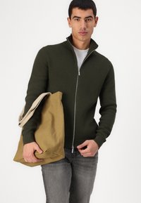 Olivengrønn zip-up genser med ribbet tekstur, kombinert med en beige lerret tote bag og grå bukser. Minimalistisk design og subtile detaljer.