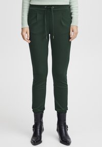 Green, fitted jogger pants with a drawstring waist, pleats, and soft texture, paired with black zippered ankle boots.