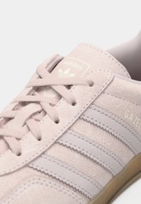 Παντόφλα από ροζ σουέτ με ανοιχτό γκρι ρίγες, στρογγυλά κορδόνια και υφασμένο καουτσούκ σόλα. Διαθέτει το "GAZE" και το λογότυπο Adidas στη γλώσσα.