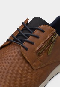 Καφέ παπούτσι με υφή, μαύρα ελαστικά κορδόνια και διακοσμητικό μπρούτζινο φερμουάρ στο πλάι κοντά στη φτέρνα.