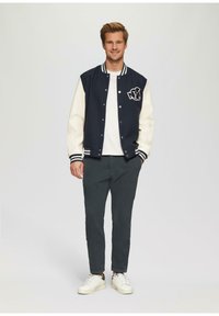 Marineblauwe en crèmekleurige varsityjas met geribbelde kraag en manchetten, voorzien van een geborduurd logo. Gekleed met donkere broek en witte sneakers.