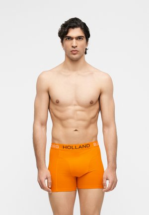 Mężczyzna bez koszulki z ciemnymi włosami, noszący jasnopomarańczowe bokserki z napisami "HOLLAND" i "BJORG" na pasku, stojący na białym tle.