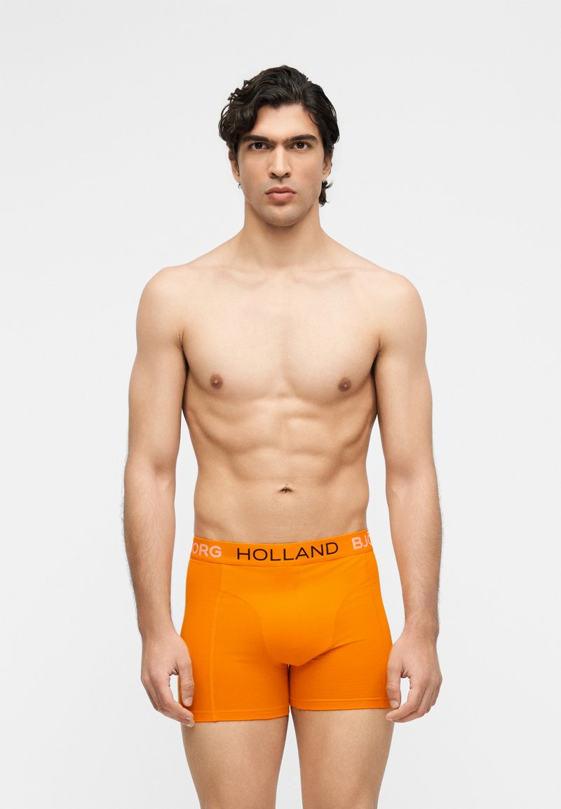 Άνδρας χωρίς πουκάμισο με σκούρα μαλλιά που φορά φωτεινά πορτοκαλί μπόξερ με τις λέξεις "HOLLAND" και "BJORG" στη μέση, που στέκεται σε λευκό φόντο.