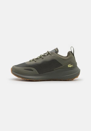 Niski sneaker w kolorze oliwkowej zieleni i ciemnoszarym z teksturowaną podeszwą, sznurowaniem z przodu, pętelką na pięcie oraz żółtym logo krokodyla Lacoste po boku.