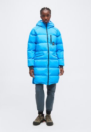 Donna che indossa un lungo piumino blu acceso con cappuccio, pantaloni grigi e scarpe da trekking marroni, in piedi davanti a uno sfondo semplice.